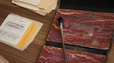 Handout A brown, long brush with a black head lying on a table. There's an old gold leaf manufacturer leaflet to the left.