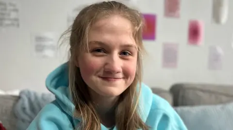 Milly is pictured in her home. She has long brown hair which is partly tied back from her face. She is wearing a turquoise hooded jumper. 