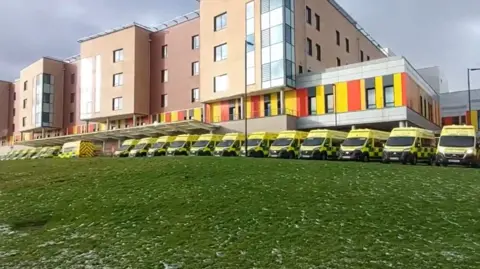 Ian Syme A row of ambulances, with the front of them visible on the photo, in the middle. The building is in the background and a large area of grass is in the bottom half of the image.