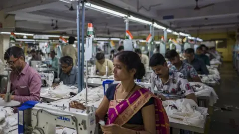 Getty Images Employees at a garment factory in India with a woman at a sewing machine in the foreground wearing a purple and gold dress