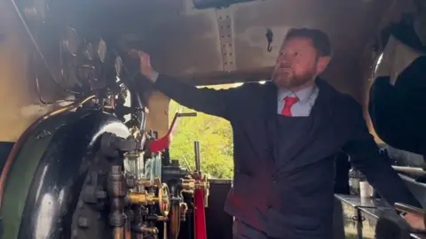 Isle of Wight Steam Railway A steam locomotive driver sounding the whistle from inside the cab. He has a beard and is wearing a blue uniform with a red tie.