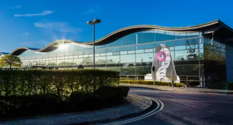 A large, glass building with a wavy roof. There is a graphic of a woman peering through one of Dyson's ring hairdryers stuck on to the right-hand side. A road with a hedge leads up to the building. 