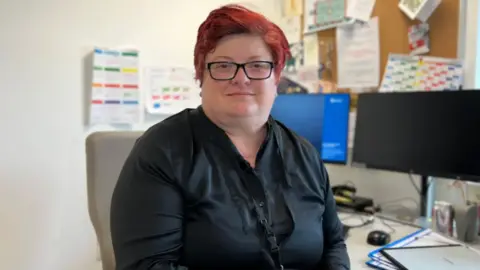 Dr Elisabet Gomes Dos Santos is sitting at her desk and is smiling at the camera. She has short red hair and is wearing a pair of glasses and a black shirt with a lanyard around her neck. There are two monitors on her desk and a few plastic files. On the wall in front of the desk is a noticeboard with various papers pinned to it and there are some pinned directly on the wall to her left.
