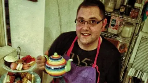 Village Bakers An old photograph of Kevin Sargeant who has dark hair and glasses and is wearing a black t-shirt and a purple apron. He is holding up a stripey teapot next to an afternoon tea stand which has a range of cakes on it