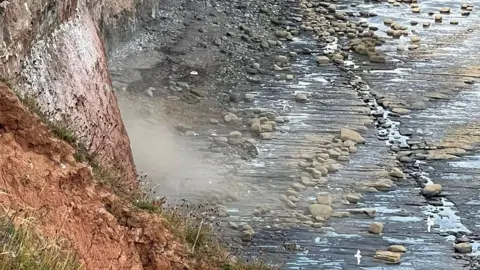 A dust cloud is billowing out from the bottom of a coastal cliff. Large rocks and pebbles are strewn across the stony beach below. Seagulls are flying above the beach.
