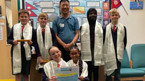 Smiling at the camera, Dawa Geljen Sherpa is surrounded by six schoolchildren. All of the pupils are wearing white Nepalese scarfs and two are holding a certificate between them. Everyone is smiling at the camera. There is schoolwork about Nepal pinned to the wall behind them.