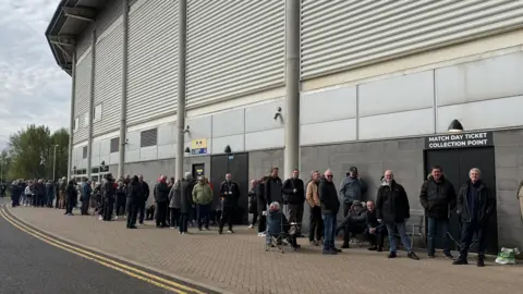 A long queue of people outside a sports stadium. They are waiting near a sign which reads “Match Day Ticket Collection Point". They are standing on a paved walkway next to a tarmac road under an overcast sky.