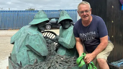 Tom Leaper crouches in front of the artwork he has made. It features two bronze fishermen holding a fishing basket. Tom himself is an older man with short grey hair and glasses, and is wearing a blue T-shirt.