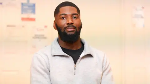 Billy has black hair, a beard and a moustache. He is wearing a white sweater with a three-quarter zip and black t-shirt underneath. He is sat in a large hall against a white wall.