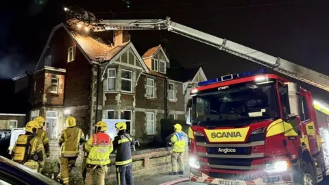 DWFRS Fire engine pulled up outside a large brick house - six firefighters are standing on the pavement in front of the building - a ladder from the fire engine stretches to its roof - it is raining.