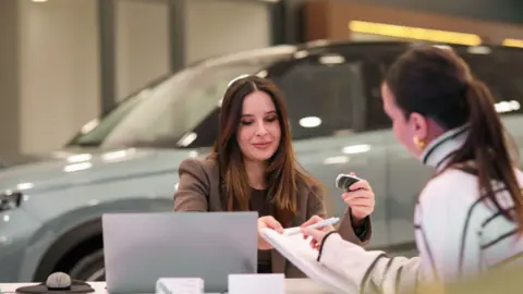 Two women signing paperwork with a car in the background