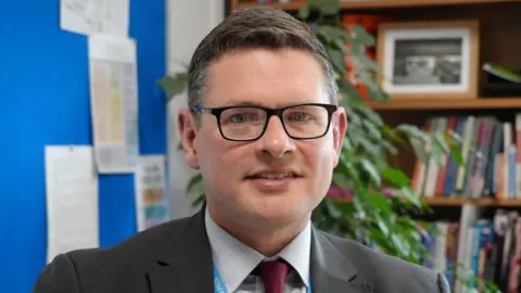 Shaun Whitmore/BBC A head and shoulders image of a man wearing glasses and a dark grey blazer over a shirt and tie. He is smiling and looking into the camera.
