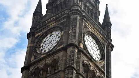 Middlesbrough Council A large tower with two clockfaces without hands visible on two sides of the building. The stone is dark grey with sets of three arches underneath the clock faces.