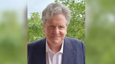Rejoin EU Party John Stevens smiles for the camera. He has thick grey hair and is wearing a blue jacket and open necked white shirt.