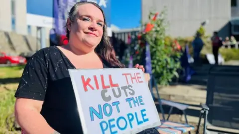 Woman holding a sign which says 'kill the cuts not the people'.