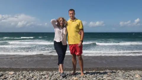 RNLI Matty and Joanna standing next to each other on Newgale beach with the sea behind them. Matty, on the right, is wearing his RNLI uniform - a yellow t-shirt and red shorts. Joanna, on the left, has long blonde hair which she is holding away from her face as the wind is blowing it. She is wearing a light pink long sleeve shirt and black trousers.