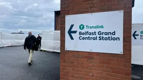 BBC A sign saying Belfast Grand Central Station in navy letters pointing to the left of a wall. There is a walkway marked out past barriers covered in white sheets.
