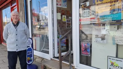 Gordon Bird A man in a grey cardigan stands proudly outside the front entrance of a newsagents