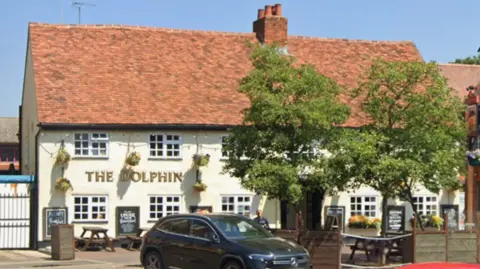 Google The exterior of The Dolphin, which is a traditional style pub. It is a wide building with 12 large windows with white grids on. There are two trees outside and several picnic tables. It has THE DOLPHIN in gold lettering on the left side, above two blackboard with chalk writing on.
