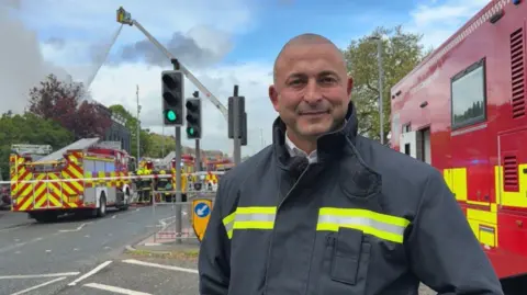 Claudia Robinson/BBC Jim Hudson smiling into the camera. He is wearing a blue jacket with a yellow hi-vis stripe across the chest. There is a red fire engine to the right. To the left, grey smoke can be seen with multiple fire engines. There is a tall crane in the middle with firefighters tackling the fire with water from it.