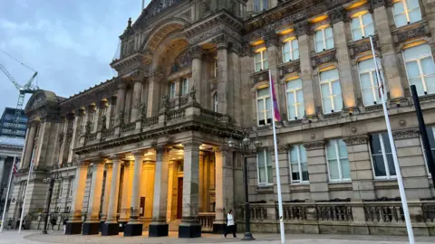 A large and imposing stone building with a columned portico at the front that is lit up, and a domed roof with four flag poles standing in front 