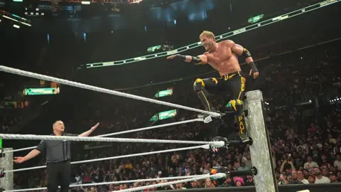 Getty Images A blonde-haired male wrestler stands poised on the top turnbuckle of a WWE ring, balancing with one foot on the ropes as he prepares to leap toward the centre of the ring. A referee stands nearby inside the ring. The arena is packed with spectators, and bright arena lights illuminate the action.