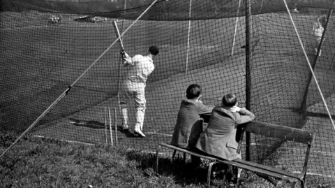 Getty Images A black and white image showing two boys sitting on a bench watching a cricketer in his whites swipe his bat at a ball. He is in nets used during training.