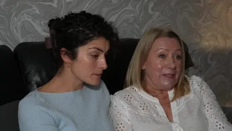 Holly Morgan, wearing a light blue jumper and her mother-in-law Jan Morgan, wearing a white top, sit on a sofa together. They both look very upset.