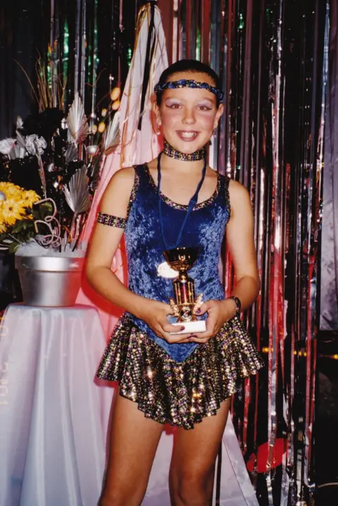 BBC / Wall to Wall Media Ltd / Richard Dowden A young girl wearing a blue and gold dancing costume and holding a gold trophy, while smiling at the camera.