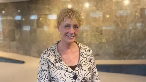 A woman wearing a white top with black flowers on, with short curly blonde hair looks at the camera