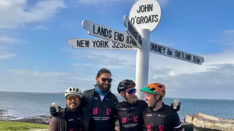 Four men including Ross MacDonald (left), and Ross MacDonald are standing in front of a white sign which says John O'Groats. The sign has several arms pointing in different directions, one of which says Lands End. The blue sea is in the background and the men are all wearing black.