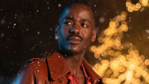 BBC Ncuti Gatwa wearing a brown jacket looking wistfully to the left of the camera with some lights flickering behind him