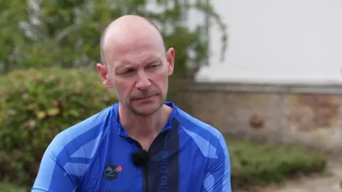 Jason Hogg, a man with a shaven head, wearing a blue cycling vest and sat in a park staring at the camera.