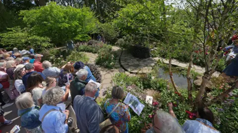 PA Media Crowd gathered around a Chelsea Flower Show garden display with stone paths, flowers, and trees on a sunny day.
