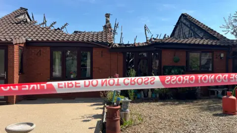 John Fairhall/BBC The outside of Siobhan Fulcher's Kenninghall bungalow. A red and white striped fire service cordon has been placed outside the property following a fire in Sunday. Part of the roof has caved in which was damaged during the blaze. The picture has been taken on a clear day with a blue sky.