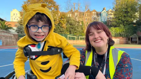 George Carden/BBC A boy with a harness to help him stand wearing glasses and a Pudsey hoodie standing next to a teacher with shoulder-length red hair and a high vis jacket