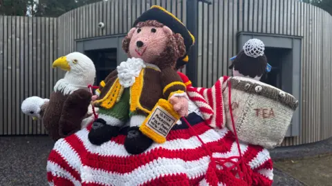 An assortment of knitted figures sits on top of a post box. A crocheted American flag lies underneath a knitted eagle, a little figure wearing old style officer clothing and a naval hat and a box of tea. It's all brightly coloured and striking.