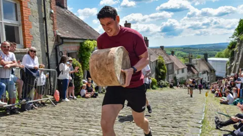 A man carrying a large wheel of cheese runs up Dorset's Gold Hill as spectators watch from the sidelines