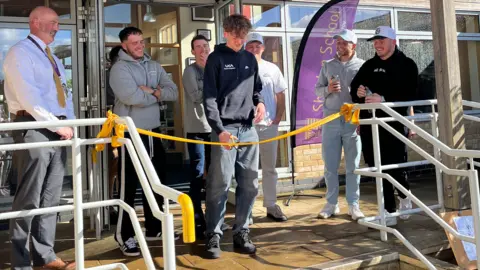 A group of people standing at the entrance to a school. One person is standing behind a yellow ribbon cutting it for the hall of fame's opening. Everyone is smiling.