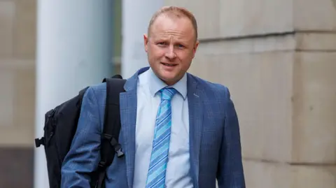 A man with fair, receding hair. He his wearing a blue plaid suit, with a light blue shirt and a tie with different colours of blue stripes. He is carrying a backpack over one shoulder. 