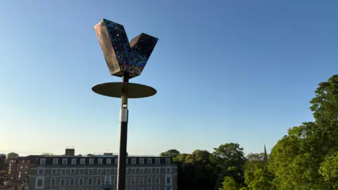 Cambridgeshire County Council The unlit beacon earlier in the day, with a view over Shire Hall. It is a metal V, with some holes in it, at the top of a pole. 