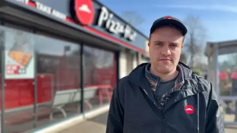 Cian standing outside a Pizza Hut franchise wearing a navy blue zip up jacket and baseball cap both with the red Pizza Hut logo on them.