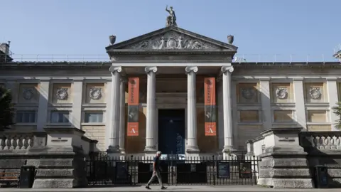 Getty Images A man walks past Oxford's Ashmolean Museum, a grand Roman-looking building with four large pillars at its front. 