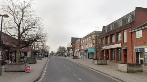 A Google street view of Victoria Road. Shops are on either side, and on the wide pavements there are large, wooden planters. All but one have thin trees in them, their branches bare.