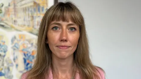 A woman with long dark-blonde hair looks at the camera. She is wearing pink lipstick and a pink jacket