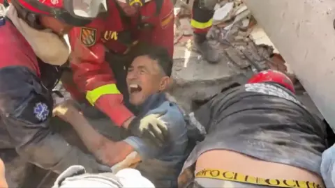 A man grinning with his mouth open amid rubble as he is rescued from a collapsed building.