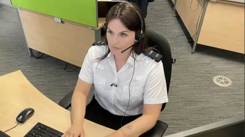 Call handler Jasmine is sat at a desk wearing police civilian uniform. She has a headset on and is looking at a computer screen. She has long dark hair worn loose and blue eyes.