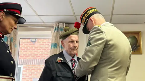 Lord Lt Jo Ropner and Brig Gen Armel Dirou pinning the insignia onto Mr Smith's lapel. They are all in military clothes.