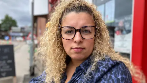 Elizabeth Baines/BBC A woman with blonde curly hair. She wears a blue leopard print shirt and large square glasses. 