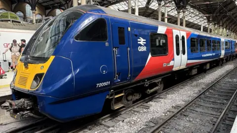 Zoie O'Brien/BBC A side view of the front of a train at Liverpool Street Station, showing the driver's cab. It is painted in bright blue with GBR written in white letters to the right of the driver's door. Beyond that there is a broad stripe of red and white across the body of the train including the first double doors for passengers. It is resting on railway tracks with more empty tracks to its right. Above it is the glass and metal roof of the station.